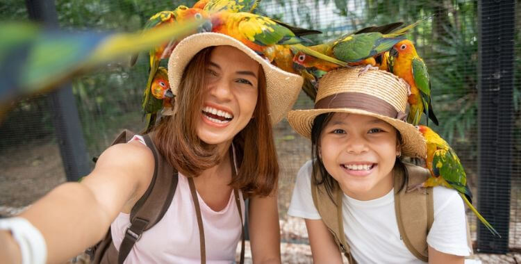 A mother and child wearing hats smiling at the zoo with parrots sitting on their hats.